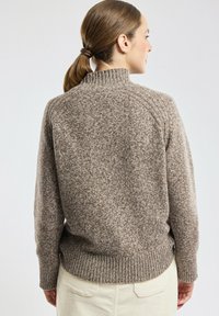 Femme aux cheveux bruns attachés, portant un épais pull en laine brun texturé et un pantalon de couleur claire, faisant face à l'opposé de la caméra.