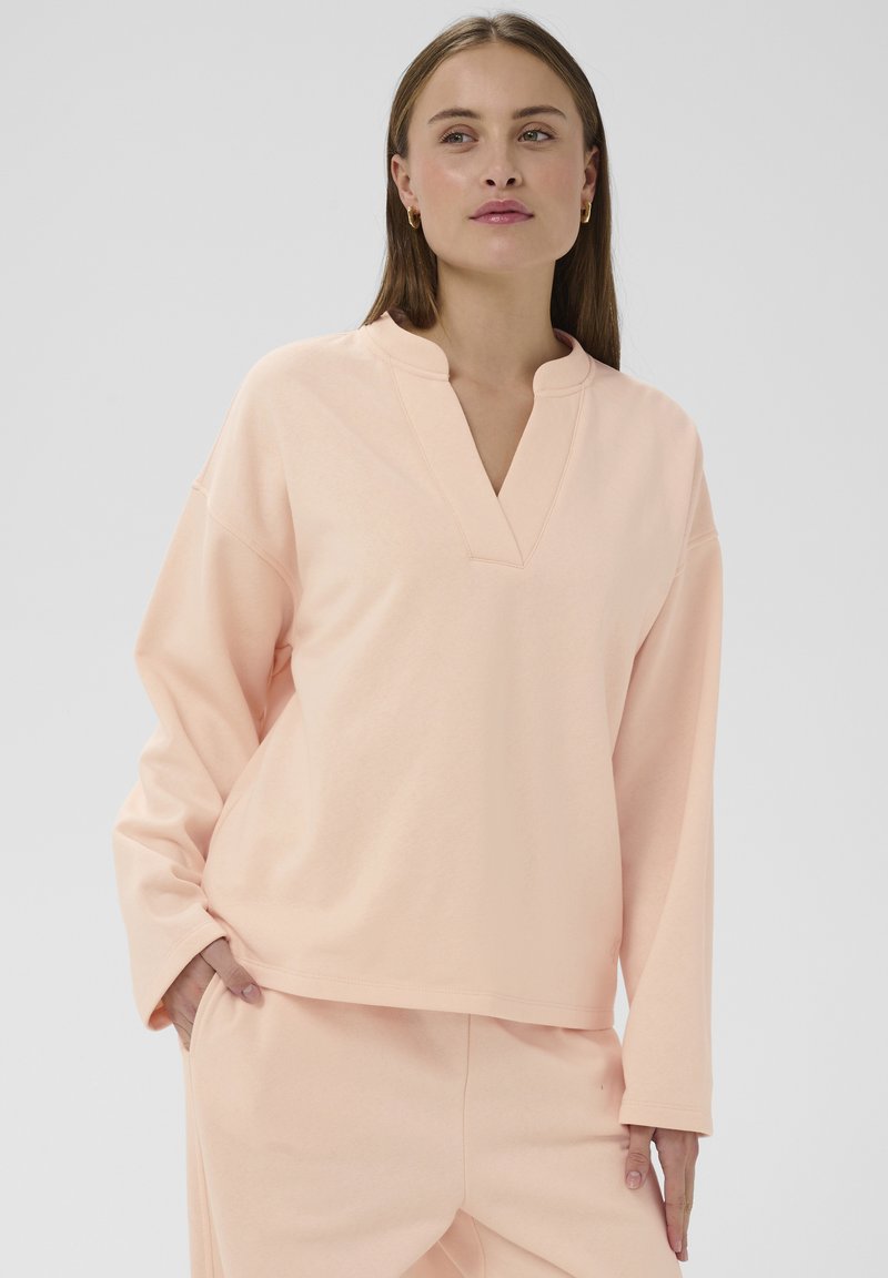 Svijetloružičasti pulover s V-izrezom i dugim rukavima. Mekani materijal s ležernim krojem i vidljivim šavovima. Nošen s odgovarajućim hlačama.