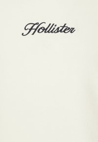 Krémová látka s vyšitým logem "Hollister" v tmavě námořnické modři. Hladká textura s jednoduchým a čistým designem.