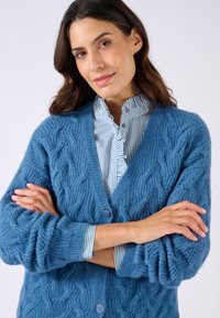 Cardigan blu in maglia con motivo a treccia, dotato di un profondo scollo a V e grandi bottoni. Sotto, una camicia a righe blu chiaro con colletto arricciato.