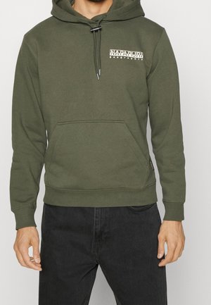 Mann trägt olivgrünen Napapijri-Hoodie mit Fronttasche und schwarze Jeans, steht vor neutralem Hintergrund.