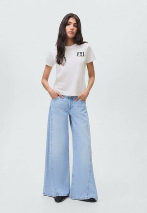 T-shirt en coton blanc avec le texte "FYI", associé à un jean en denim large de couleur bleu clair. Tissu doux et coupe décontractée avec des détails de couture visibles.