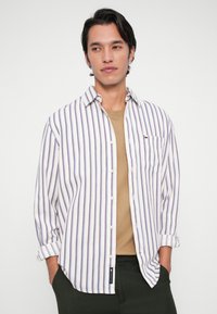 Νεαρός άνδρας με σκούρα μαλλιά, φορώντας ένα λευκό και καφέ ριγωτό πουκάμισο πάνω από ένα μπεζ μπλουζάκι, με τα χέρια στις τσέπες των σκούρων παντελονιών του.