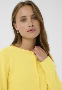 Cardigan jaune en maille douce avec un col rond et six boutons assortis, présentant un tissu texturé et des poignets côtelés.