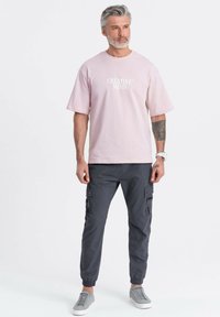 Muž se šedými vlasy a vousy, oblečený v světle růžovém tričku "KREATIVNÍ MYŠLENÍ", tmavě šedých cargo kalhotách, šedých teniskách a bílém náramkovém hodinách.