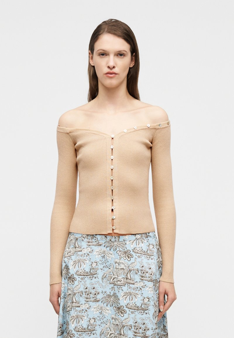 Γυναίκα με ίσια καστανά μαλλιά φοράει μπεζ ριμπ  πουκαμίσα με ξεκούμπωτα κουμπιά και γαλάζια φούστα με ανθισμένο σχέδιο, στέκεται μπροστά σε άσπρο φόντο.