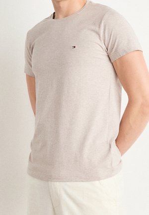 Homme portant un T-shirt beige à manches courtes avec un petit logo rouge, blanc et bleu sur la poitrine et un pantalon de couleur claire.