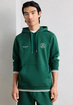 Nuori mies, joka pukeutunut metsänvihreään huppariin, jossa on valkoinen reunus, etutasku ja tekstilliset logot rinnassa, seisoo yksinkertaisen vaalean seinän edessä.