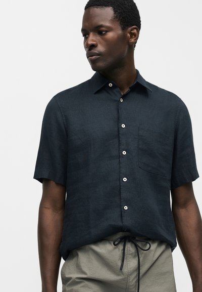 Άνδρας με σκούρο μπλε πουκάμισο με κοντά μανίκια και τσέπη στο στήθος και μπεζ παντελόνι με κορδόνι, κοιτάζει αριστερά σε μονόχρωμο φόντο.