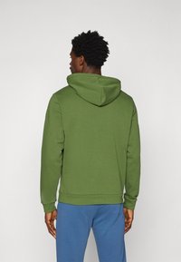 Grønn hettegenser laget av mykt stoff, med kengurulomme og ribbede mansjetter. Kombinert med blå joggebukser.