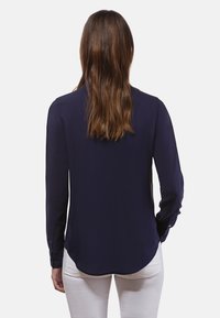 Marineblaues Langarmhemd mit abgerundetem Saum und geknöpften Manschetten, aus glattem Stoff, von hinten betrachtet. Getragen mit weißen Hosen.