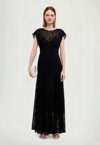 Μαύρο maxi φόρεμα από δαντέλα με εφαρμοστό τοπ και φουσκωτό φούστα, διαθέτει κοντά μανίκια και διάφανο κορσέ από δαντέλα. Το μοντέλο κρατά μια μικρή λευκή τσάντα.