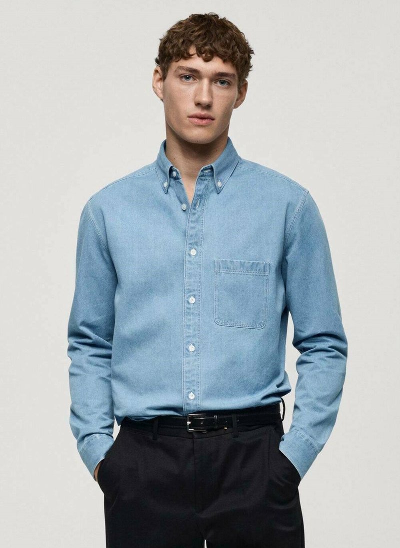 Camicia in denim azzurro chiaro con bottoni, taschino sul petto, maniche lunghe e una texture liscia, abbinata a pantaloni scuri.