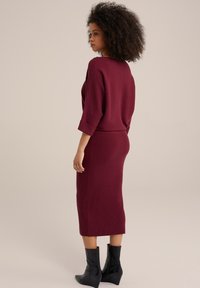 Bordo pleteni dvoslojni outfit koji se sastoji od lepršave majice s 3/4 rukavima i uske midi suknje. Tekstuirana tkanina i crne čizme do gležnja.