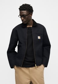 Moški, ki nosi črno jakno na zadrgo z logotipom Carhartt, črno majico, bež hlače in okrogla očala, roke v žepih, pogleda usmerjenega navzdol levo.