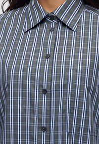 Gros plan d'une personne portant une chemise boutonnée avec des rayures verticales et horizontales bleues, blanches et grises et un col pointu.