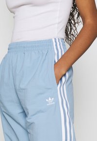 Person som bär ljusblå Adidas träningsbyxor med vita ränder och logotyp, hand vilande i fickan, ihop med en vit ärmlös topp.