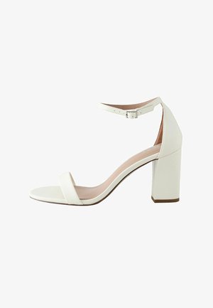 Hvid højhælet sandal med ankelrem og spænde, åben tå og blokhæl på en ensfarvet hvid baggrund.