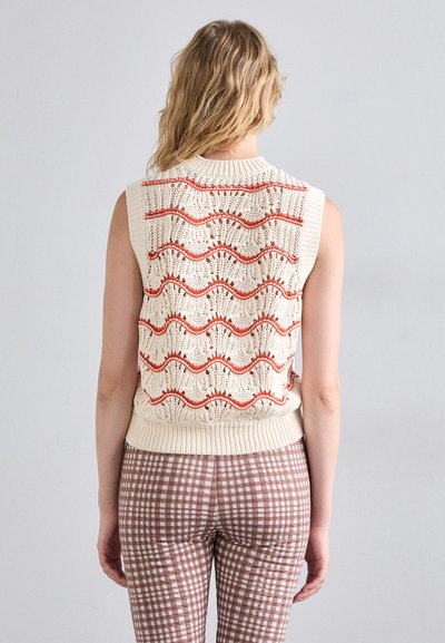 Haut sans manches en maille crème avec des motifs rouges ondulés ; présente un col et un ourlet côtelés. Associé à un pantalon marron à carreaux.