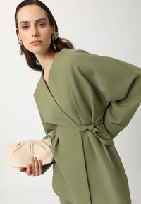 Olive green wrap dress with oversized sleeves, side tie detail; holding a light beige, gathered clutch bag. Silk-like texture, minimalistic design.