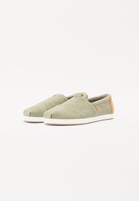 Zapatillas de lona verde sin cordones con punta redonda, acento de talón naranja en contraste y suela de goma blanca. Tejido texturizado con detalles cosidos.