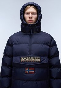 Giacca puffer blu navy con colletto alto, cappuccio regolabile, patch con logo e dettaglio della bandiera norvegese sul davanti. Texture liscia, design voluminoso.