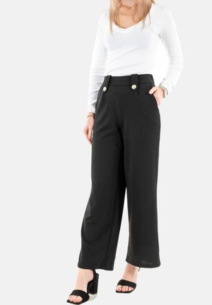 Pantalons larges noirs avec poches avant et accents de boutons dorés, associés à un haut blanc à manches longues ajusté et des sandales à talons carrés noires.