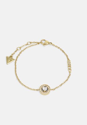 Zlata verižica za zapestje s centralnim okroglim obeskom, ki ima prozoren kamen v obliki srca, obdan s številnimi malimi prozornimi kamni in oznako z logotipom.