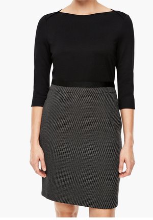 Kobieta ubrana w czarną bluzkę z rękawami do łokci oraz wysoką taliowaną spódnicę w czarno-biały wzór, stojąca z ramionami swobodnie opuszczonymi.