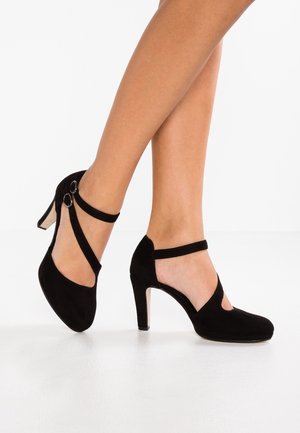 Sorte ruskind højhælede sko med udskåret design og krydsede ankelstropper. Har en spids hæl og glat tekstur.