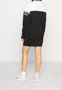 Person som bär en svart och vit långärmad sweatshirtklänning med vita sneakers, står mot en neutral vit bakgrund, sett bakifrån.