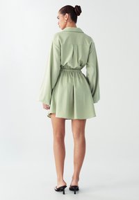 Šviesiai žalias sukriošias su ilgomis rankovėmis, elastinga juosmens dalimi ir platėjančia sijone. Lygus audinys, iki kelių ilgio, dėvimas su juodais aukštakulniai sandalais.