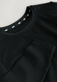 Cămașă neagră cu mâneci lungi, cu guler rotund, buzunar frontal și textură netedă. Eticheta din interiorul gulerului poartă cuvântul „self.”