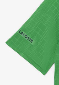 Tela verde con un patrón geométrico, que presenta un logo de Lacoste en azul oscuro sobre un parche texturizado. Textura suave y diseño de manga corta.