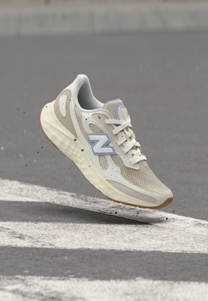 Lichtbeige en witte hardloopschoen met mesh- en suède-details, zwevend boven een weg met witte verkeersstrepen, merklogo aan de zijkant.