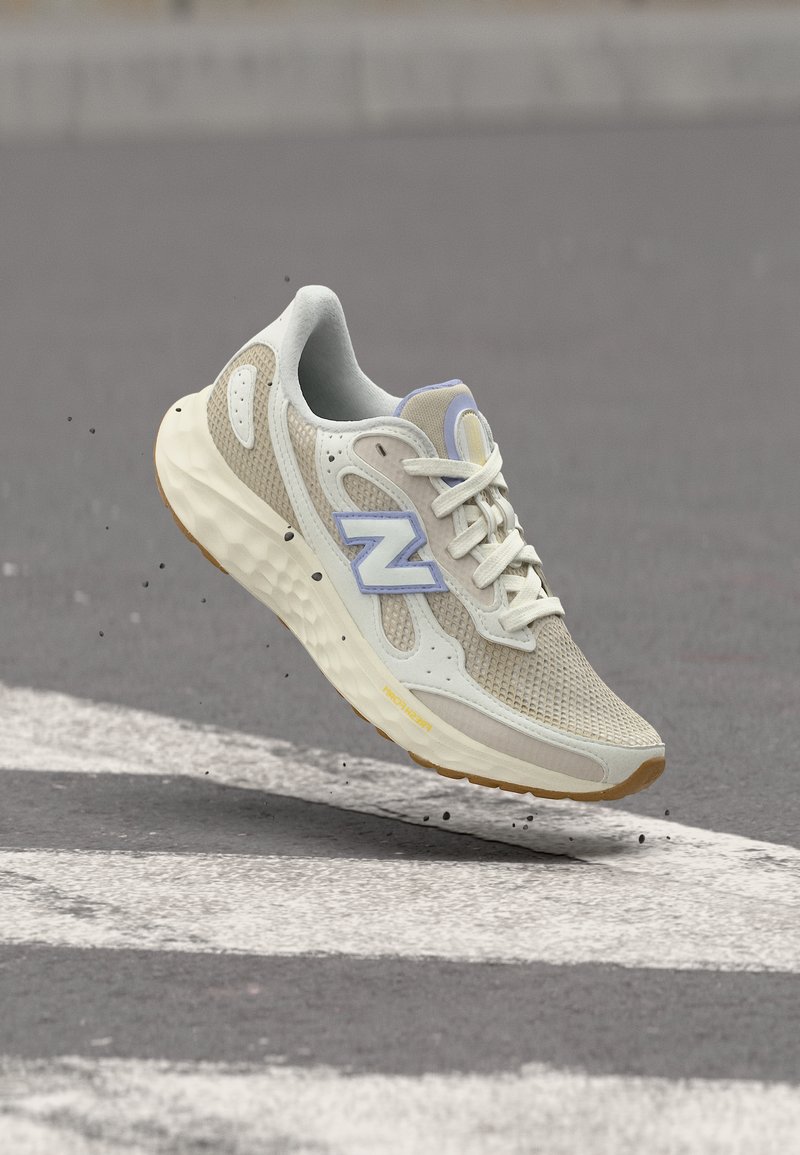 Ανοιχτό μπεζ και λευκό αθλητικό παπούτσι με λεπτομέρειες από δίχτυ και σουέντ, στον αέρα πάνω από δρόμο με λευκές βαμμένες γραμμές, λογότυπο της εταιρείας στην πλευρά.