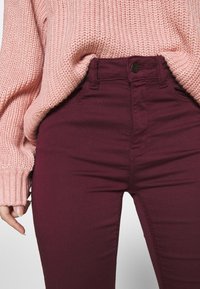 Burgundowe spodnie skinny z wysokim stanem, wykonane z gładkiego materiału, z przednimi kieszeniami, zamkiem błyskawicznym i metalowym zapięciem.