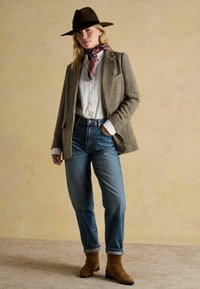 Hnedý plsťový klobúk, kockovaný sako, biela košeľa, vzorovaný šál, džínsy s vysokým pásom a hnedé semišové členkové topánky. Textúrované neutrálné pozadie.