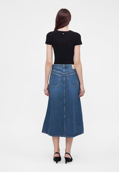 Femme aux longs cheveux bruns portant un haut noir à manches courtes, une jupe midi en jean bleu avec poches arrière et des chaussures noires à talons, vue de dos.