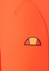 Jasnopomarańczowa tkanina z zakrzywionym logo obrysowanym na czarno, z żółtym środkiem i dwoma czerwonymi kształtami po bokach, z wyraźnym ściegiem wzdłuż szwu.