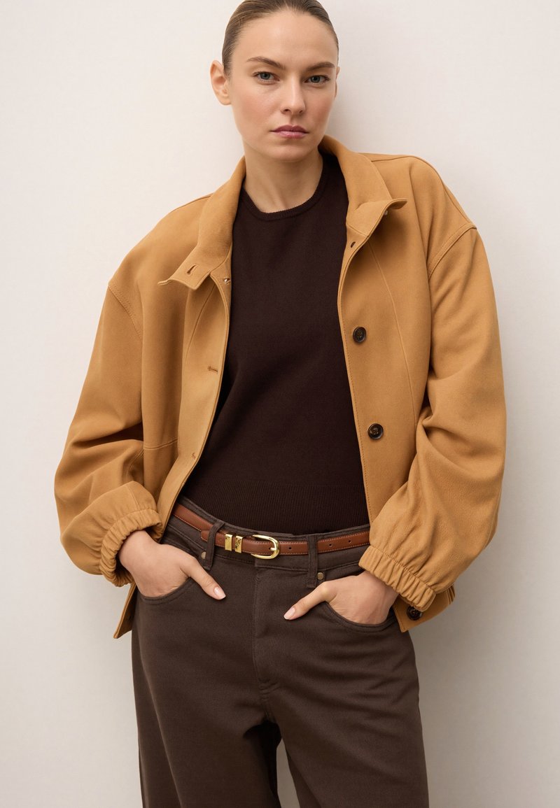 Brūns žakets ar brīvu piegriezumu, kas aprīkots ar apkakli un pogām ar spiedienu. Valkāts virs tumša džempera un apvienots ar brūnām biksēm un jostu.