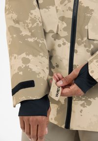 Beige waterdichte jas met een spetterpatroon. Voorzien van een rits aan de voorkant, een zwart klittenbandaccent bij de mouw en een label met de tekst "W-PRO."