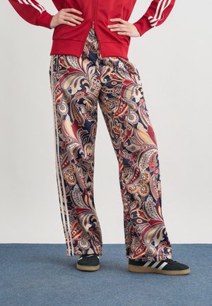 Persona che indossa una giacca sportiva rossa e pantaloni a gamba larga con motivo paisley colorato e strisce laterali, in piedi su un tappeto blu.