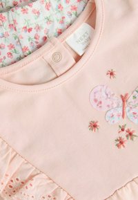 Rosa Babytop mit gesticktem Design von Schmetterlingen und Blumen, weichem Stoff, Rüschensaum und Druckknopfverschluss am Hals.