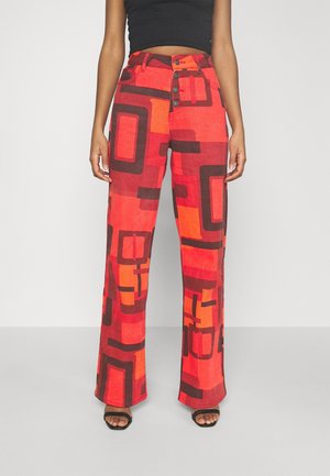 Kobieta ubrana w spodnie z wysokim stanem i szerokimi nogawkami w odważne czerwono-pomarańczowe geometryczne wzory oraz czarne sandały, w połączeniu z czarnym crop topem.