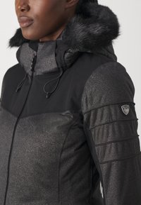 Mustahdud musta takki, jossa on kiiltävä tekstuuripinta, keinoturkistakki, vetoketjusuljenta ja pehmustetut olkapäädetailit. Logopatchi hihassa.