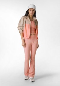 Lichte, kleurgebroken jack met een beige bovenkant en koraal onderkant, gecombineerd met een bijpassende koraalkleurige crop top en uitlopende broeken. Witte pet en sneakers.