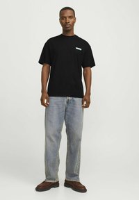 La camiseta de algodón negra presenta mangas cortas y un pequeño gráfico en color teal en el pecho, emparejada con unos vaqueros de denim azul de corte holgado y zapatos marrones.