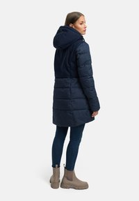 Giacca puffer blu navy con cappuccio, caratterizzata da una texture trapuntata e un tessuto a contrasto nella parte superiore. Abbinata a jeans scuri e stivaletti beige.