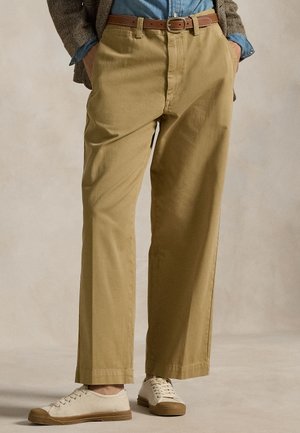 Person trägt beige gerade geschnittene Hose, braunen Gürtel, Jeanshemd, Fischgrät-Blazer und weiße Leinwand-Sneaker, Hände in den Taschen.
